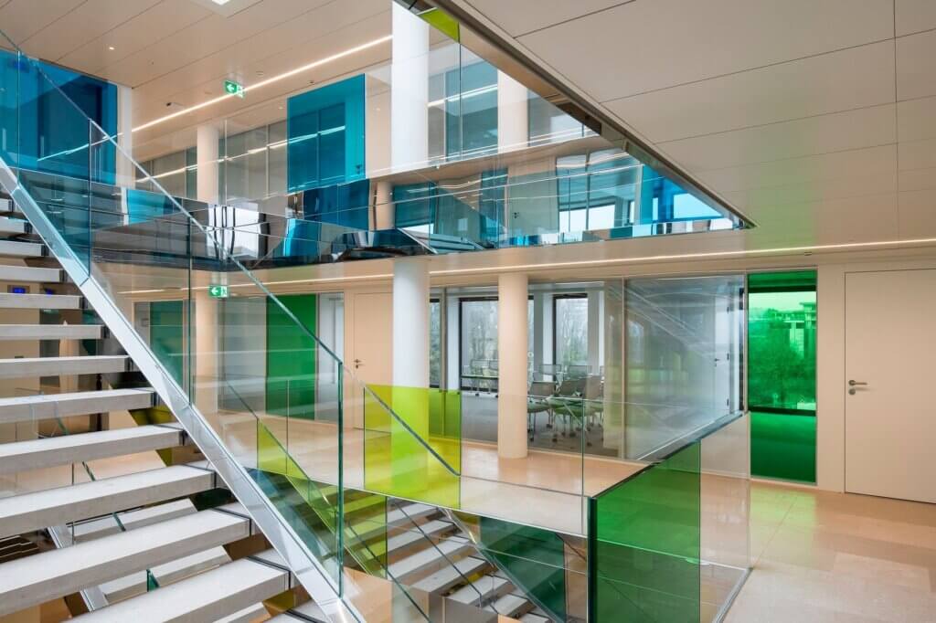 Eine moderne Büroeinrichtung mit Glaswänden und farbigen Glaspaneelen in Blau, Grün und Gelb. Eine offene Treppe mit Glasgeländer verbindet mehrere Etagen. Große Fenster lassen natürliches Licht herein. Ein Konferenzraum ist sichtbar.