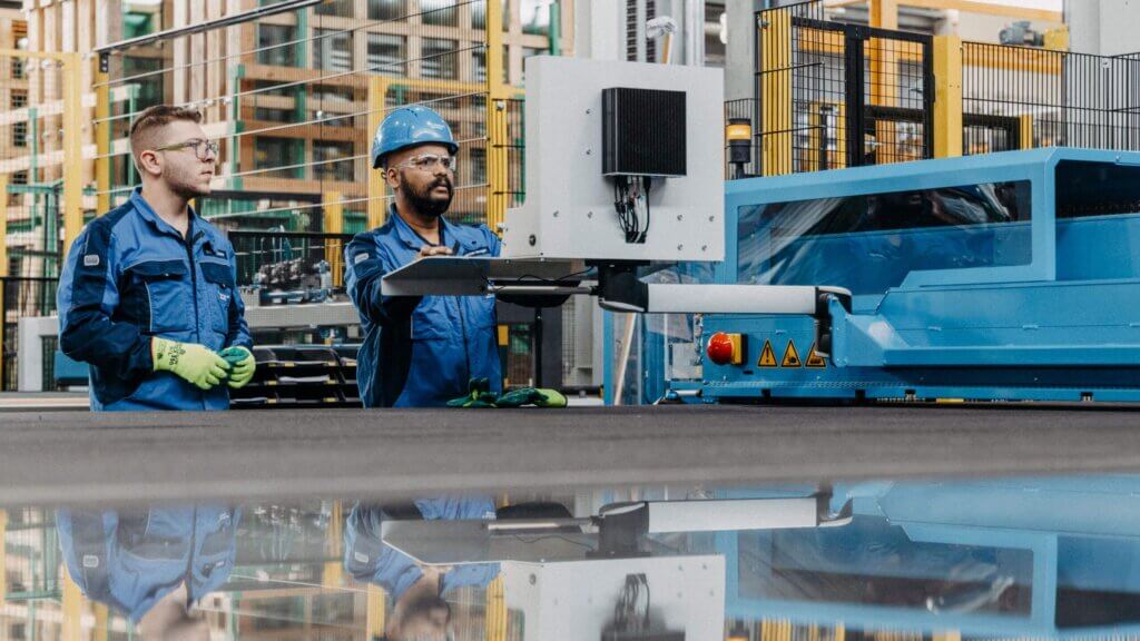 Due operai in uniforme blu e attrezzatura di sicurezza manovrano un grande macchinario industriale con un pannello di controllo in un moderno impianto di produzione.