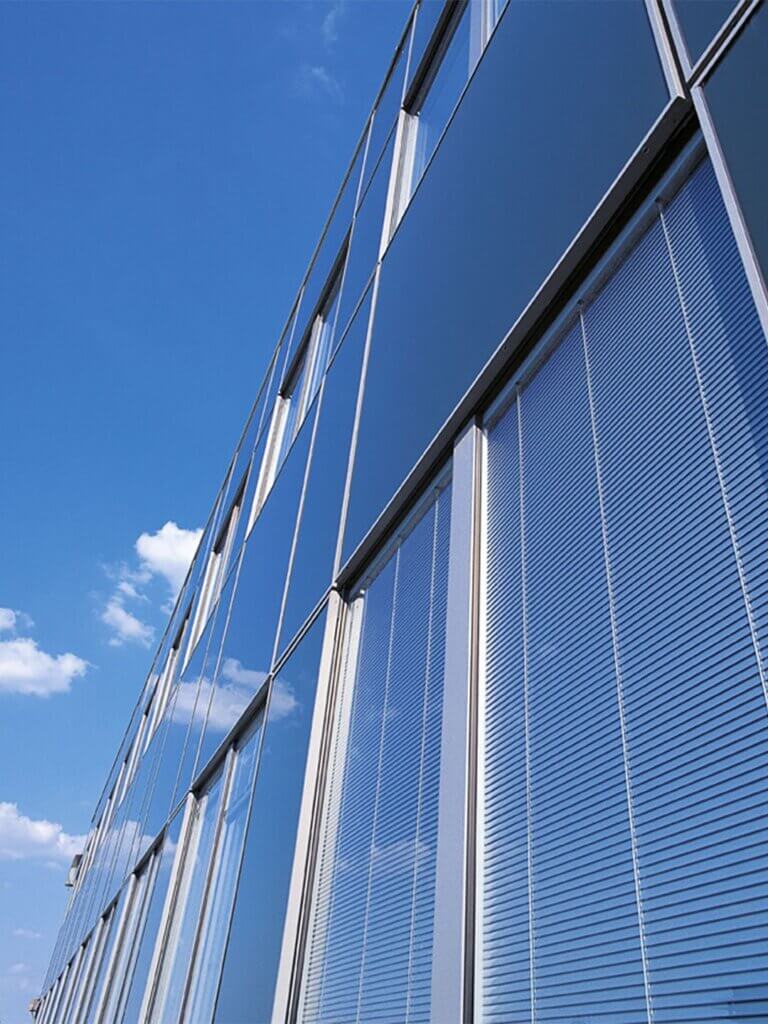 Ein modernes Glasgebäude mit geschlossenen Jalousien spiegelt einen klaren blauen Himmel mit ein paar vereinzelten Wolken. Das Foto wurde aus einem niedrigen Winkel aufgenommen, wodurch die klaren Linien und spiegelnden Oberflächen des Gebäudes betont werden.