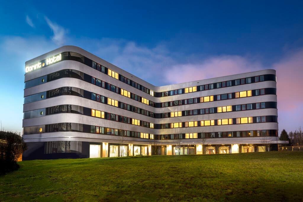 Un hôtel moderne de plusieurs étages, aux bords incurvés et aux nombreuses fenêtres éclairées de l'intérieur, se dresse dans un ciel crépusculaire. L'enseigne de l'hôtel est illuminée et le bâtiment est entouré d'une pelouse bien entretenue.
