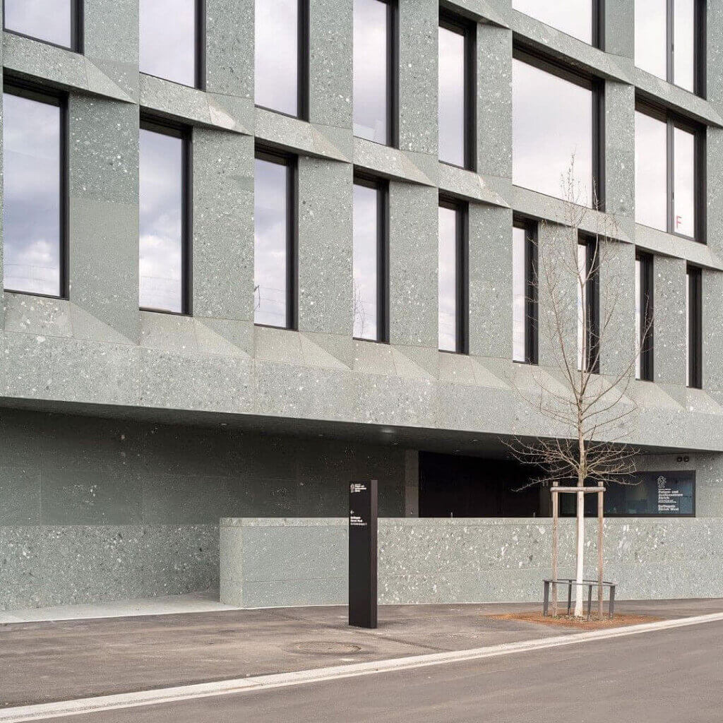 Ein modernes Gebäude mit großen rechteckigen Fenstern und einer Fassade aus blassem grünlich-grauem Stein. Davor stehen ein blattloser Baum, der von Pfählen gestützt wird, und ein großes schwarzes Schild auf dem gepflasterten Gehweg.