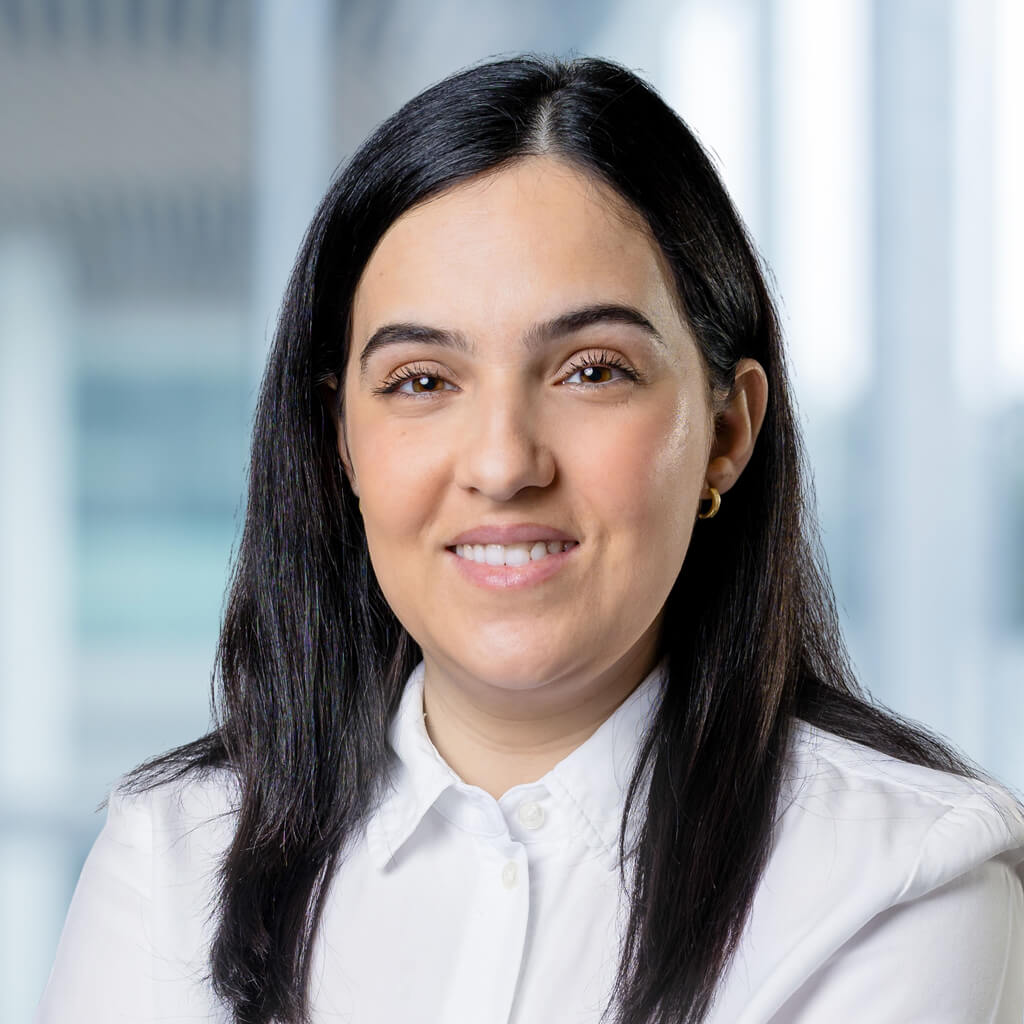 Eine Frau mit glattem schwarzem Haar, die ein weißes Button-up-Hemd trägt und sanft lächelt, posiert vor einem unscharfen Bürohintergrund mit großen Fenstern.