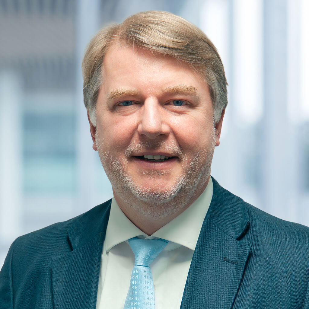 Un uomo di mezza età con capelli chiari e barba corta indossa un abito blu, una camicia bianca e una cravatta azzurra e si trova di fronte a uno sfondo sfocato di un ufficio. Sorride leggermente.